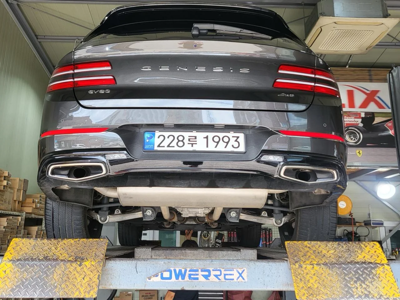The 2023 Genesis GV80 raised on a two-post lift in a Korean workshop, seen from behind, with the exhaust and rear suspension visible underneath.