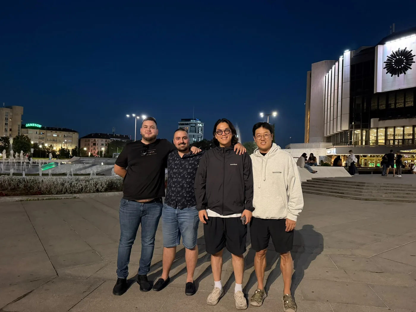 Four men standing together at night on the plaza in front of the National Palace of Culture in Sofia, Bulgaria, with fountains behind them.